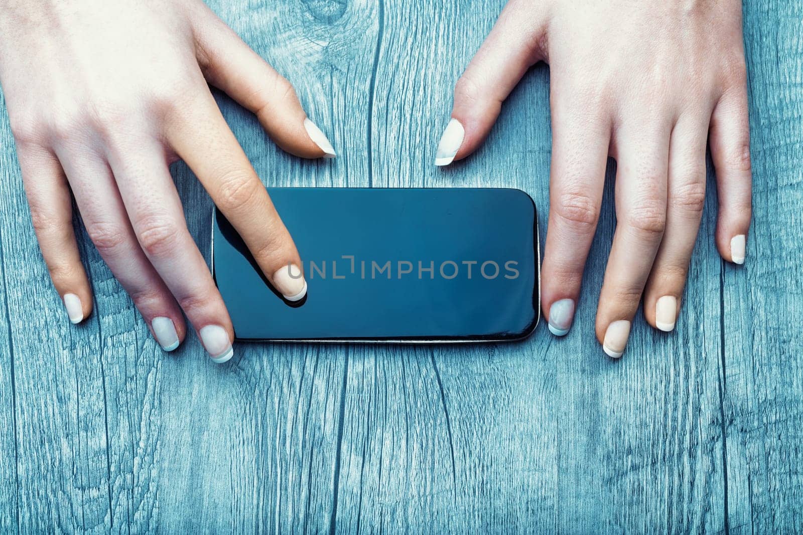 smartphone lies on a wooden surface, bordered by two hands with light skin and manicured nails. The images soft lighting and textured background suggest minimalism and calm