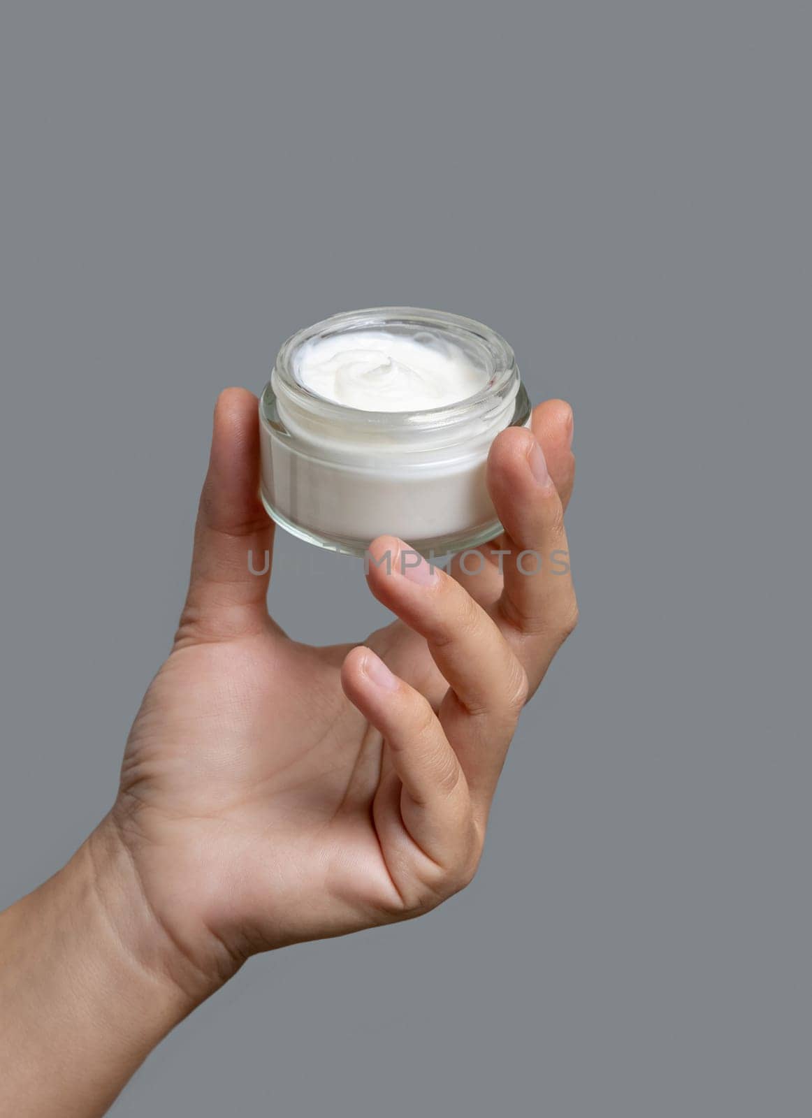 Female hand holding cream jar against grey background close up. Natural product for facial care, everyday skincare routine. Opened cosmetic Container. Spa and Wellness concept