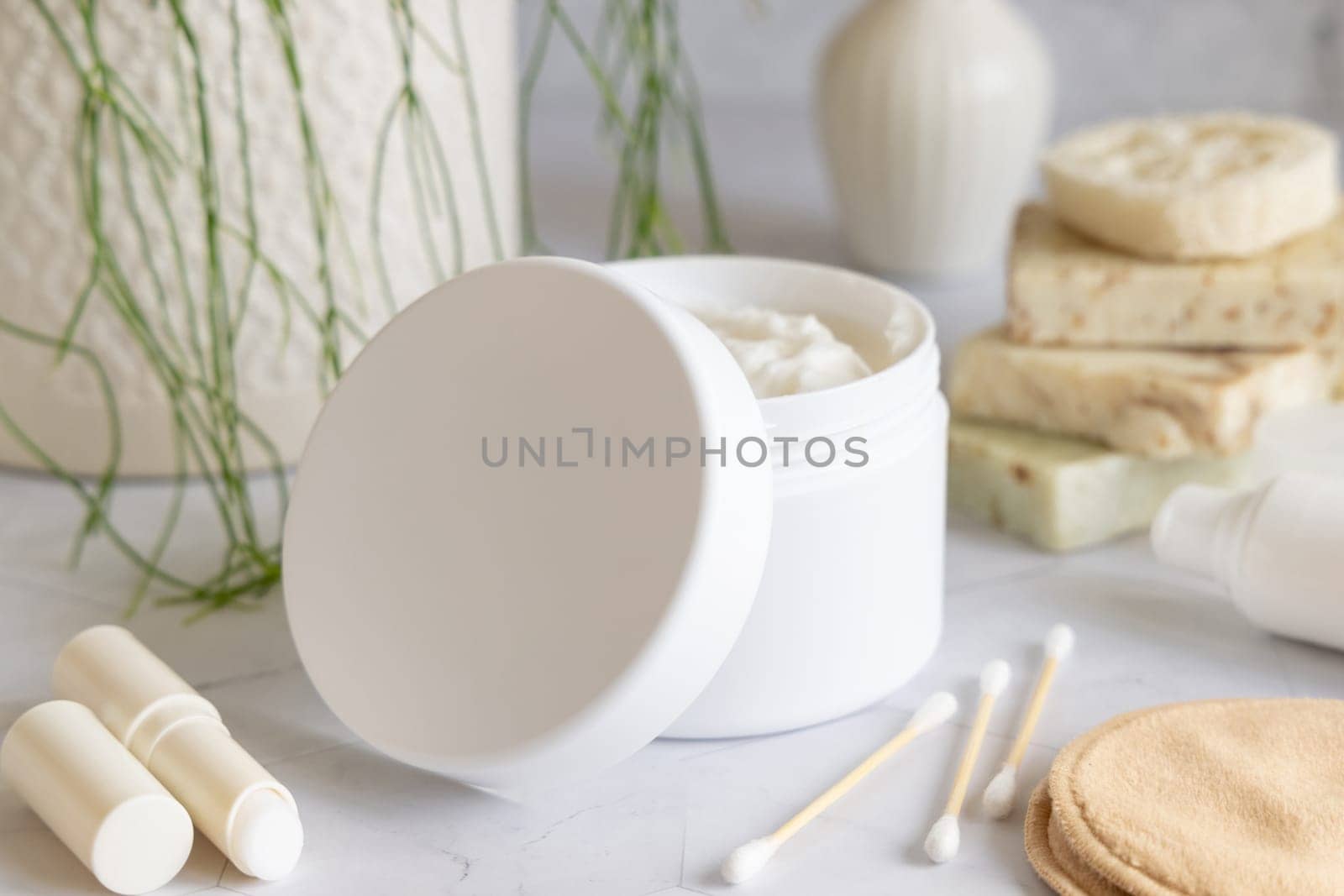 Opened cream jar with white blank lid on white marble against potted green plant and soap bars in bathroom, close up, packaging mockup. Cosmetic product for bodycare, beauty routine