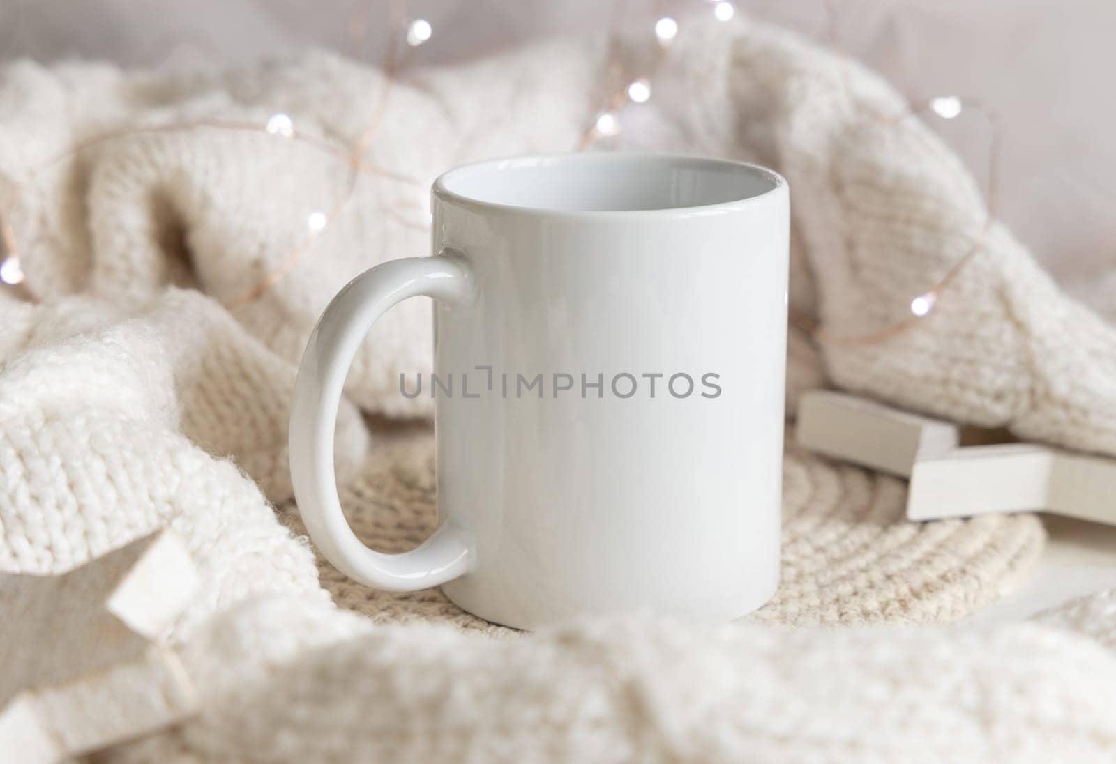 White blank coffee mug near white knitted sweater, wooden Christmas decor and lights, copy space. Winter atmospheric mock up in rustic or scandinavian style