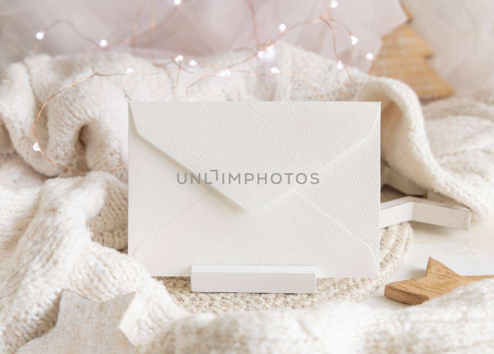 White blank envelope near white knitted sweater, wooden Christmas decor and lights, copy space. Winter atmospheric mock up, family greeting in rustic or scandinavian style