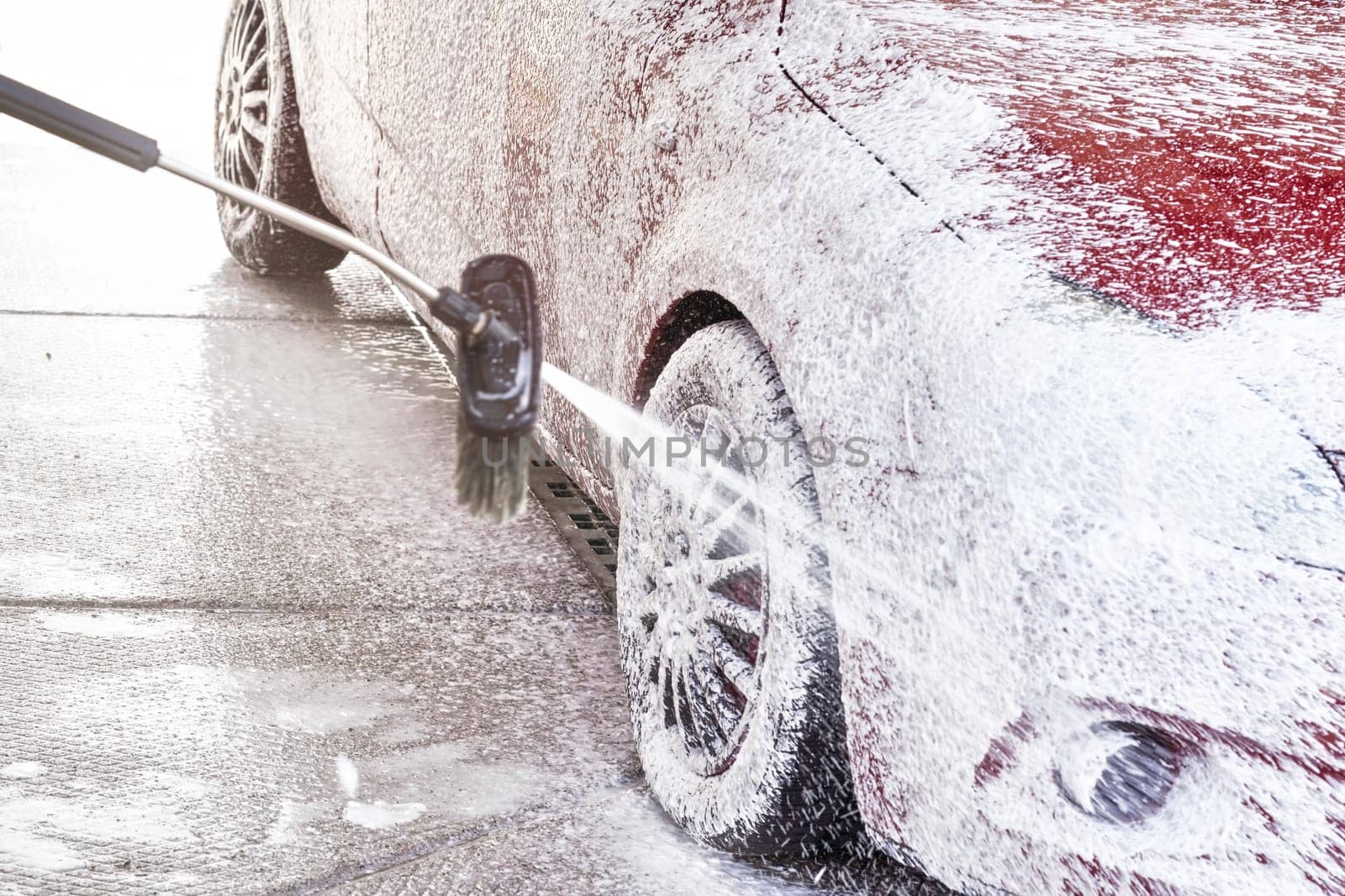 Red car washed in self serve carwash, detail on white soap foam spraying from high pressure hose onto wheel