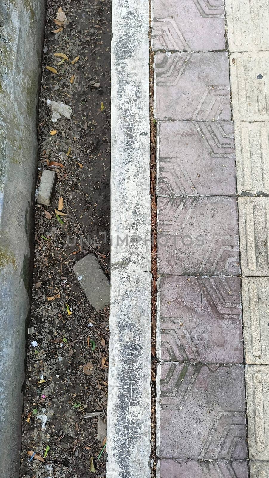 An urban scene showing the contrast between a patterned sidewalk and a dirt pathway. The textured surfaces highlight urban decay and natural elements coexisting in a city environment.