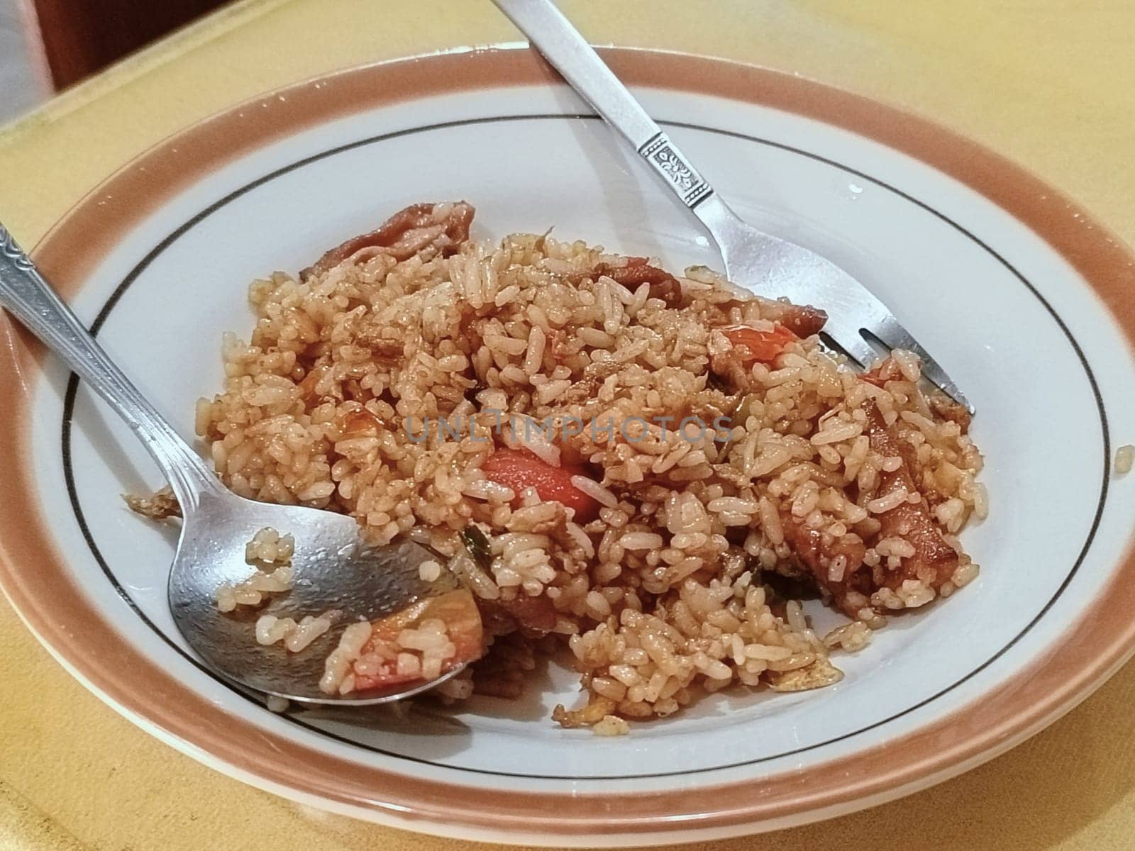 A plate of flavorful fried rice is served with a spoon and a fork on a wooden table. The dish includes pieces of vegetables and meats, showcasing a homemade and appetizing meal.