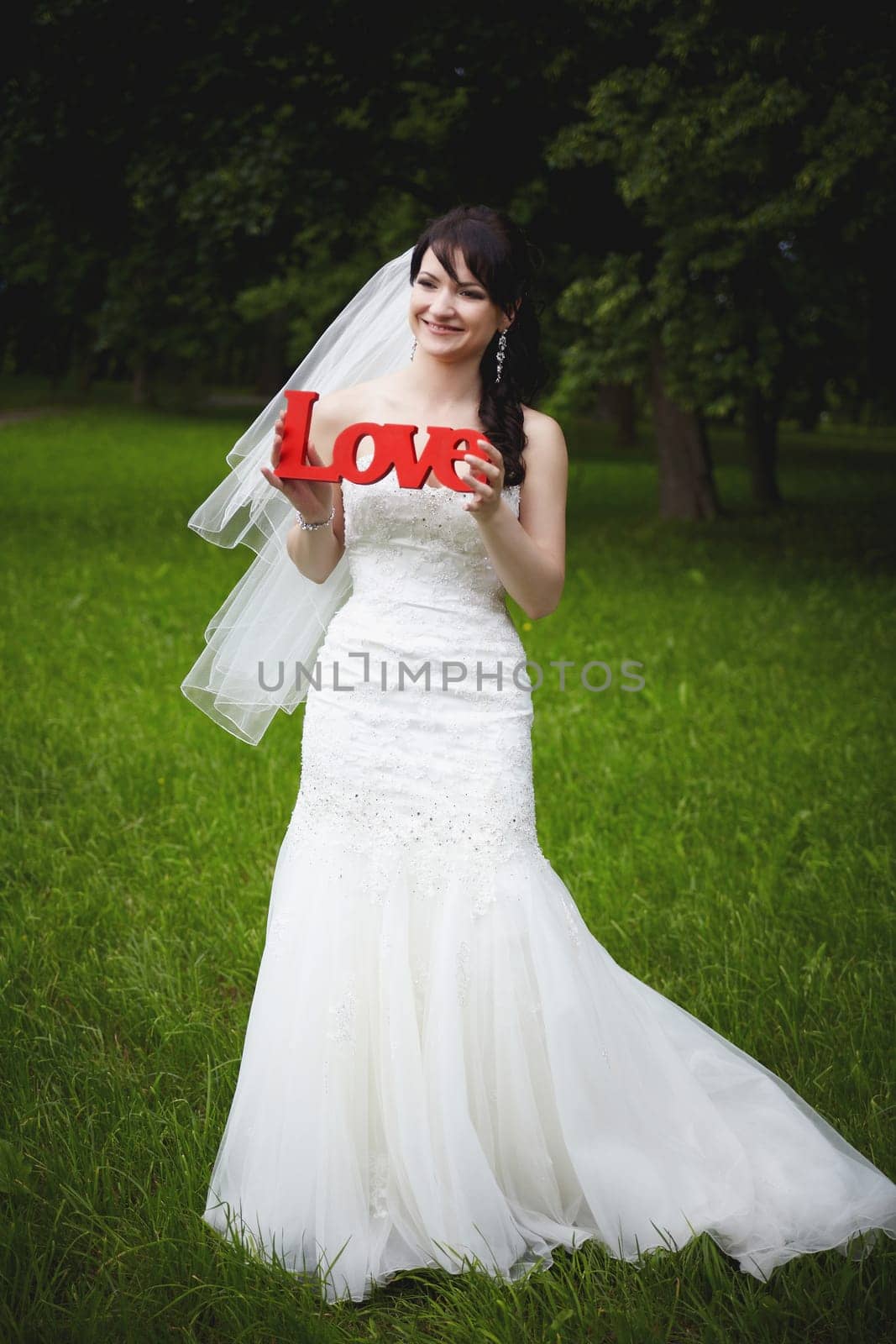 A joyful bride, dressed in a stunning dress, holds a sign of love in her hands, surrounded by lush, bright greenery