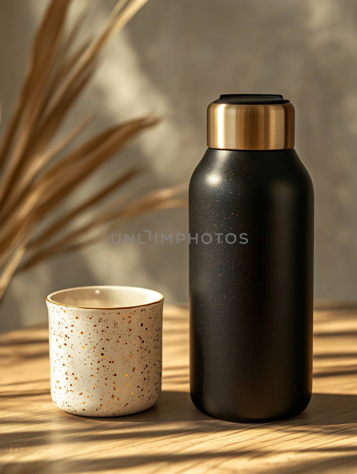 A sleek black water bottle stands next to a speckled cup on a sunlit table, showcasing a minimalist design in warm tones.