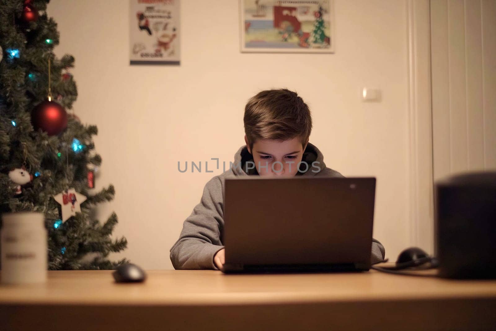 A young boy sits at a wooden table, intently working on a laptop, surrounded by holiday decorations and a Christmas tree.