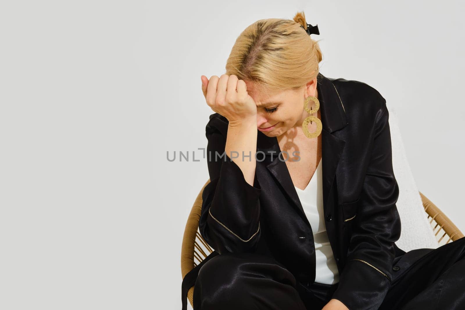 A woman in a black outfit sits in a wicker chair, her head down as she appears deep in thought. The serene atmosphere suggests introspection or contemplation in a minimalist setting.