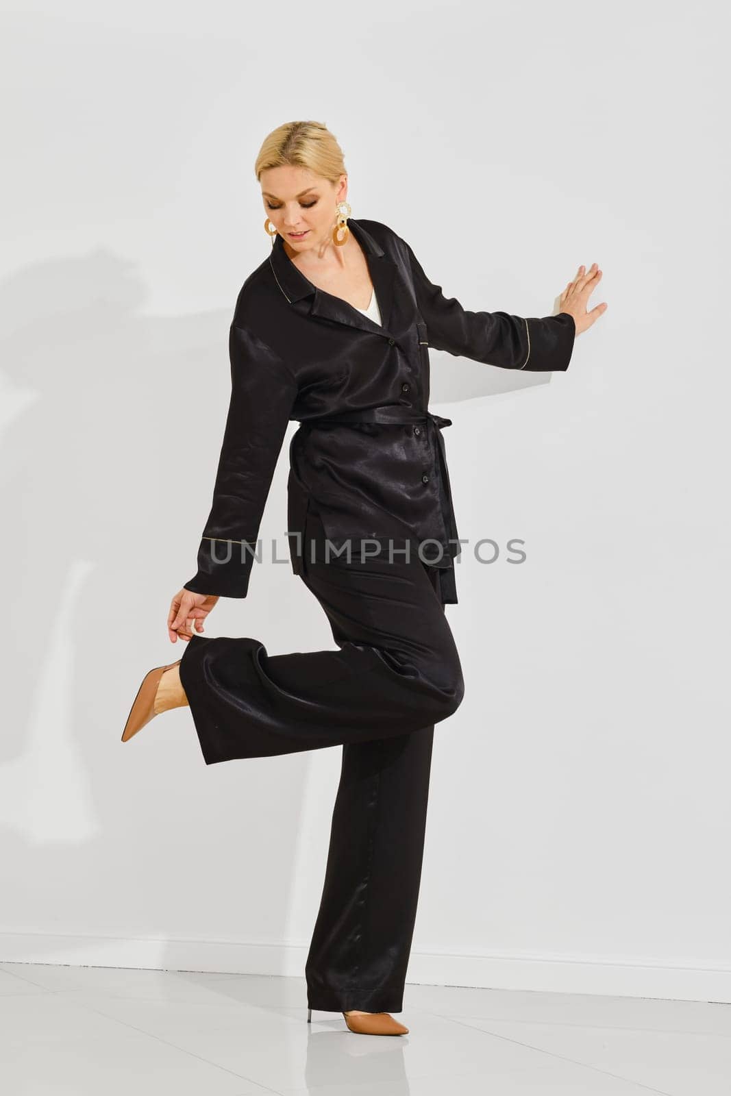 Woman in black loungewear touching the heel of her shoe, posing thoughtfully against a clean white background. Her poised posture and fashionable outfit highlight modern style and comfort.