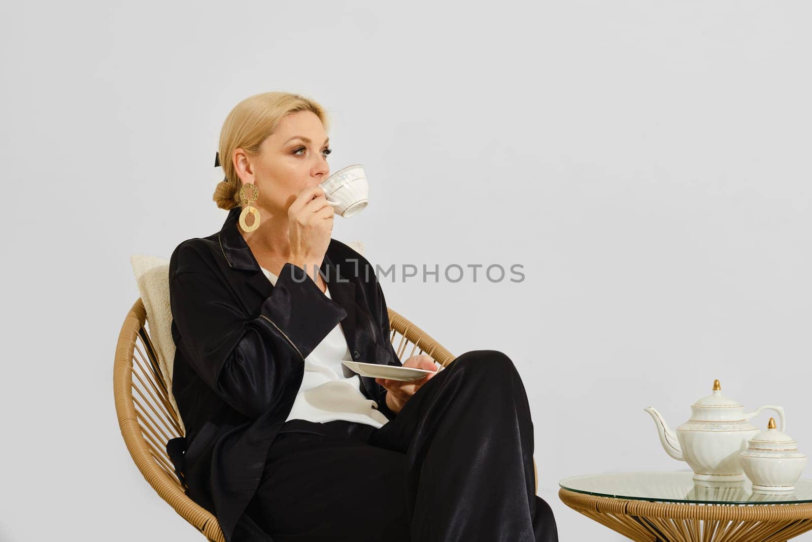 A woman is peacefully sipping tea from a delicate cup while sitting comfortably on a rattan chair. She is dressed in a casual outfit, creating a relaxed atmosphere in a minimalist room.