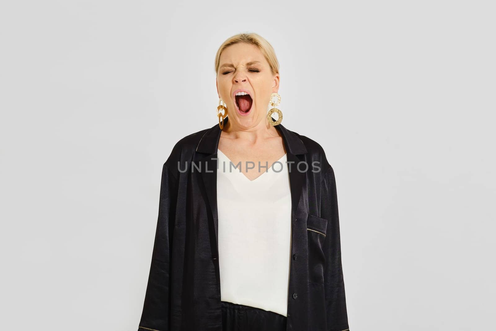 A woman stands in a minimalist studio and yawns, showcasing her relaxed demeanor. She wears a simple top and black outerwear, accessorized with bold earrings, highlighting a casual yet stylish look.