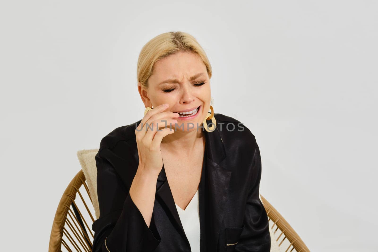 Crying woman with blonde hair sits in a stylish chair, tearing up and expressing deep emotions.