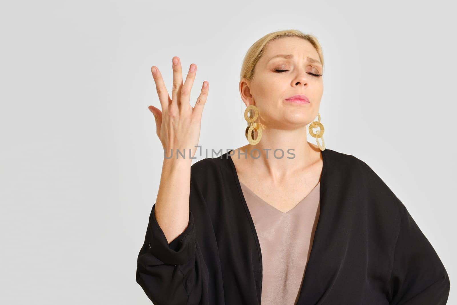 A woman stands with her eyes closed and hand raised in a gesture of frustration. She wears elegant earrings and a fashionable outfit, clearly conveying her feelings in a minimalist setting.