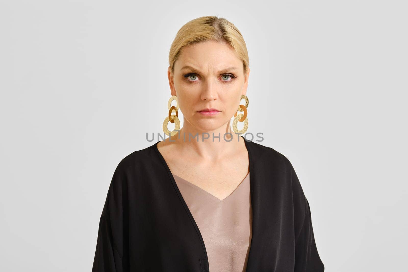 A woman stands tall with a serious expression, wearing large gold earrings and a dark outfit. Her blond hair is styled elegantly, creating a striking contrast with the simple background.
