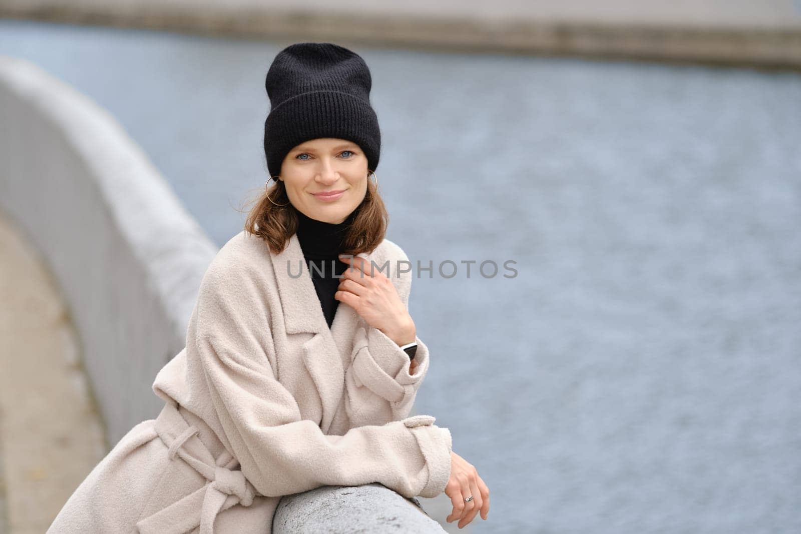 A joyful woman enjoys a peaceful moment by the riverbank, wearing a warm beige coat and a black beanie, exuding a relaxed vibe on a cool day. The gentle water reflects her calm demeanor.