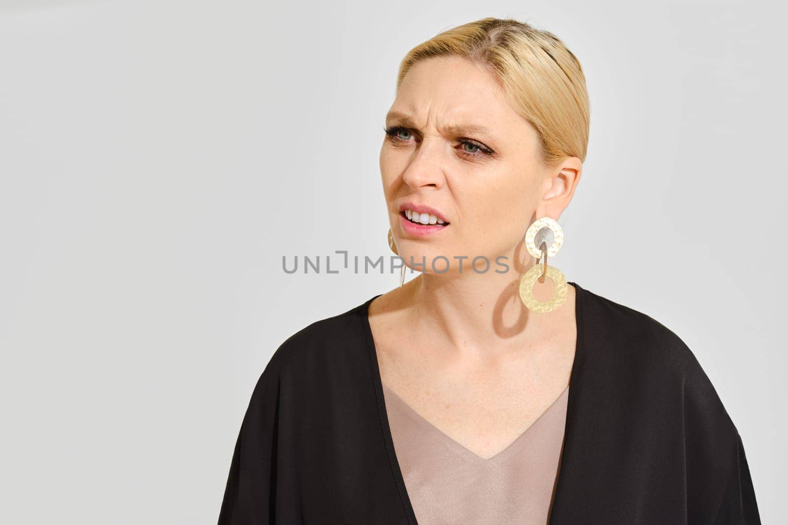 A woman in a black outfit stands against a light gray background, displaying a confused expression. Her large statement earrings and hairstyle emphasize her thoughtful demeanor, creating contrast.