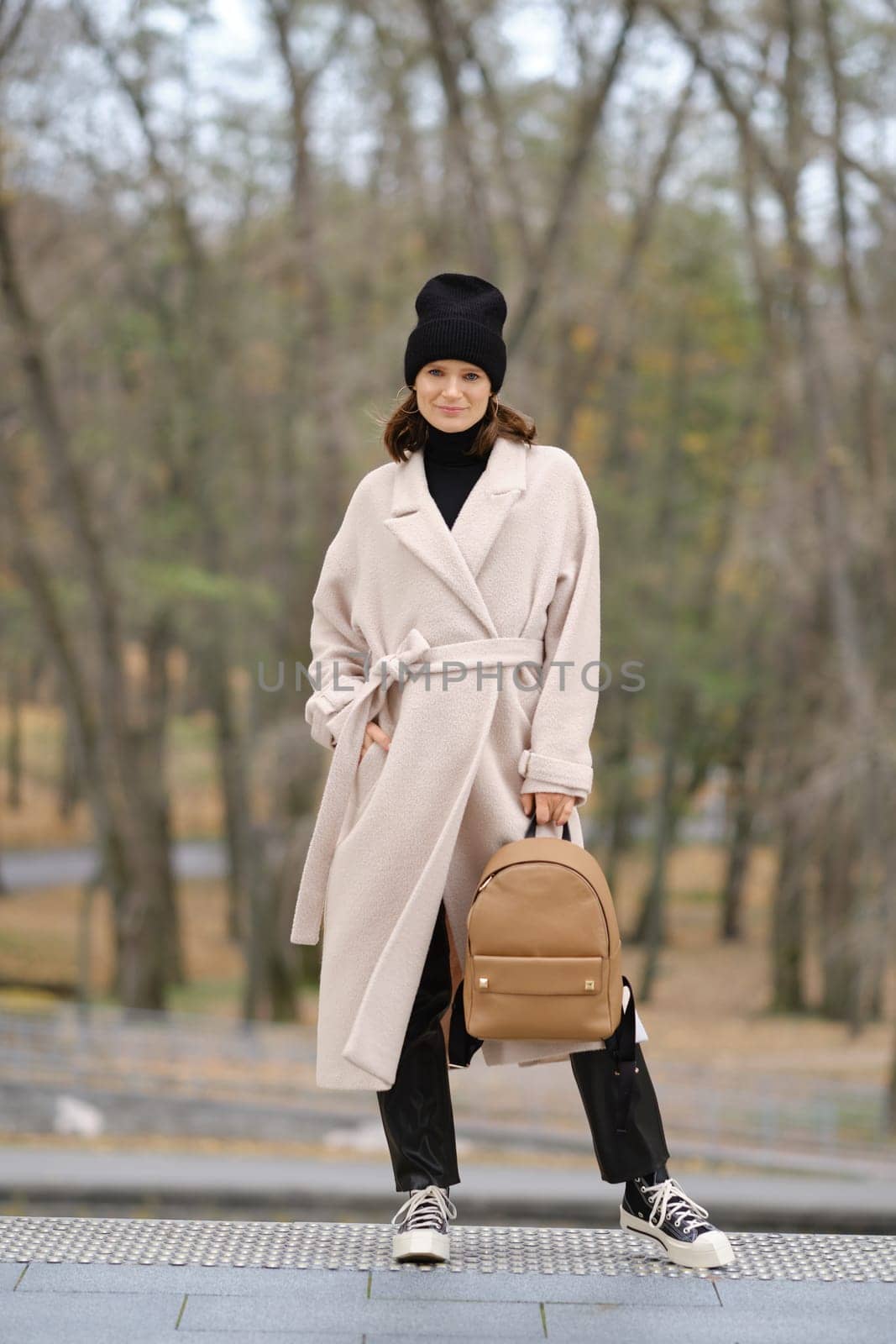 A fashionable woman stands confidently in a light coat and black beanie, holding a stylish backpack. The backdrop features a serene forest with faint autumn colors, creating a calm atmosphere.
