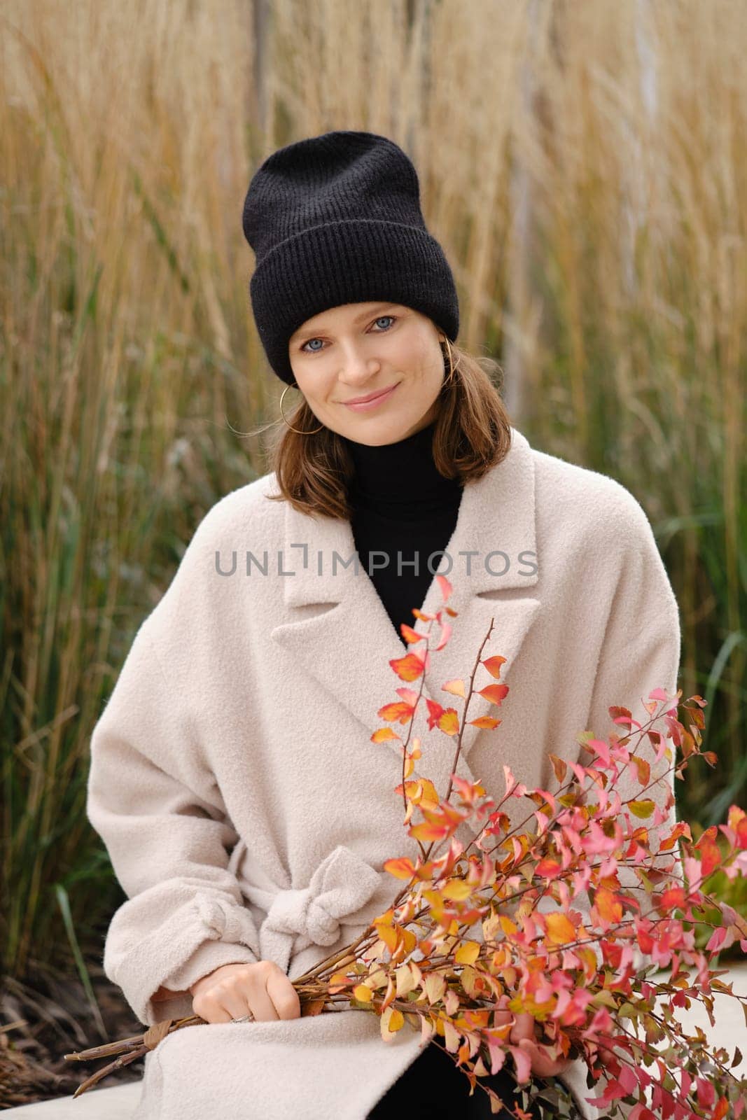 A cheerful woman sits peacefully surrounded by tall grasses, wearing a soft beige coat and black beanie.