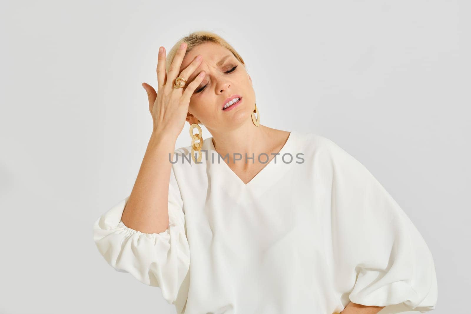 A woman is standing in a minimalist studio with a neutral background, expressing signs of stress as she lightly touches her forehead.