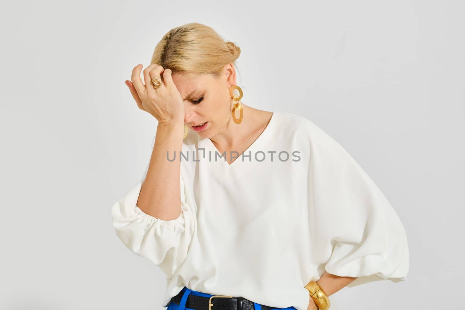 A woman with blonde hair wearing a white blouse stands with her hand on her forehead, visibly frustrated. Her expression conveys a sense of concern or disappointment in a neutral setting.