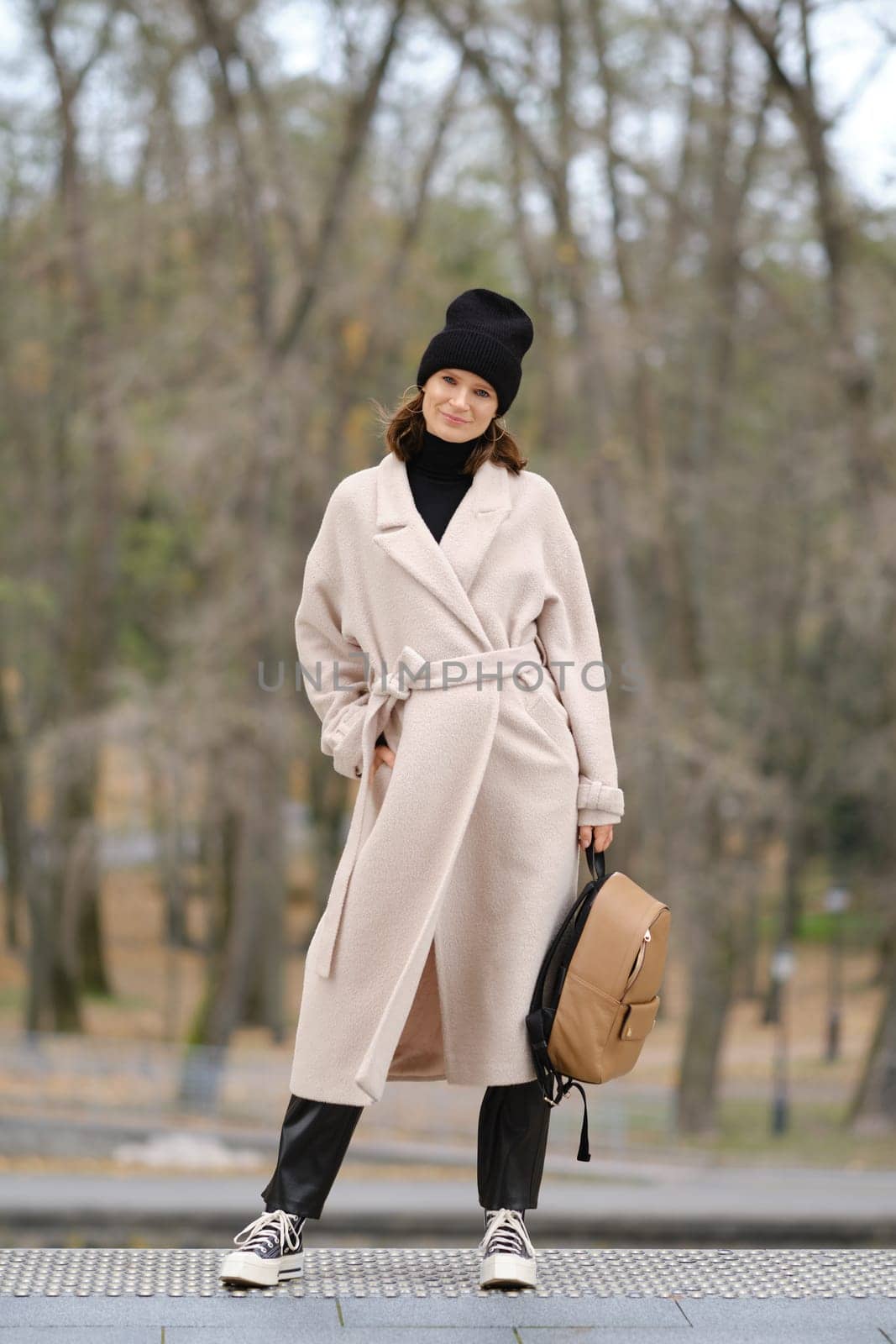 A woman dressed in a beige coat and black beanie poses confidently in a serene park setting. The trees show signs of autumn, creating a warm, inviting atmosphere while she carries a backpack.