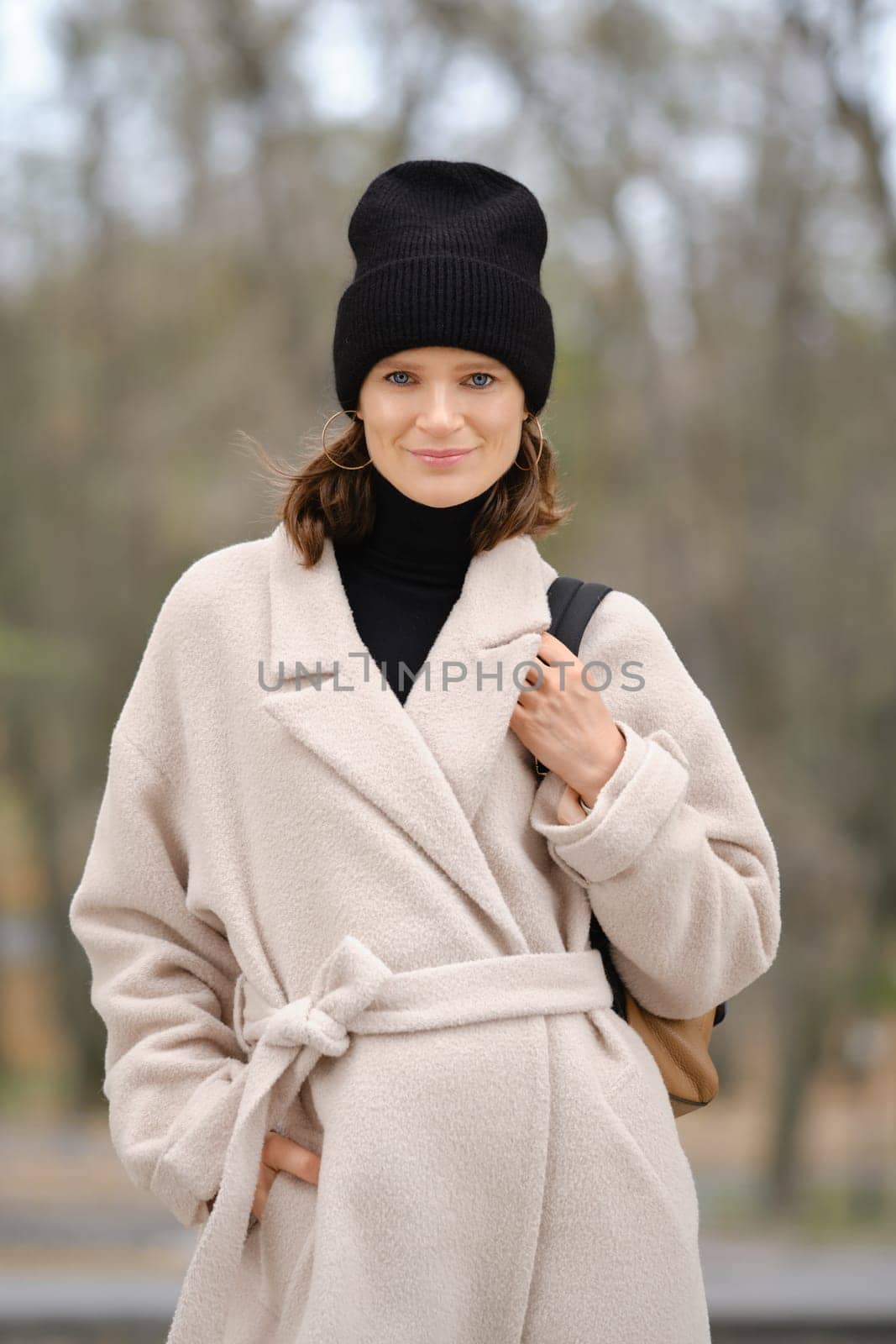 A woman wearing a stylish beige coat and a black beanie stands outdoors, smiling gently while enjoying the winter atmosphere. The blurred background suggests a calm, chilly day perfect for a walk.