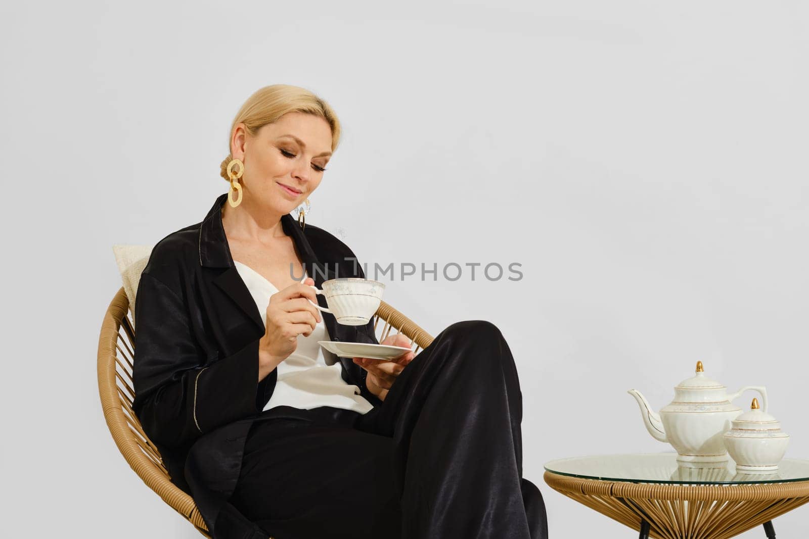 A woman in stylish attire savors a cup of tea, sitting comfortably in a chic chair. The serene atmosphere highlights the beauty of the tea set beside her, emphasizing relaxation and sophistication.
