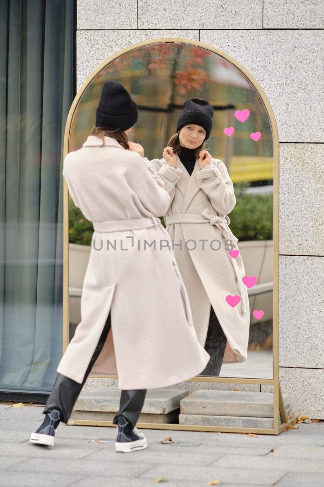 A woman stands outdoors, looking at her reflection in a modern mirror while wearing a coat and a beanie.