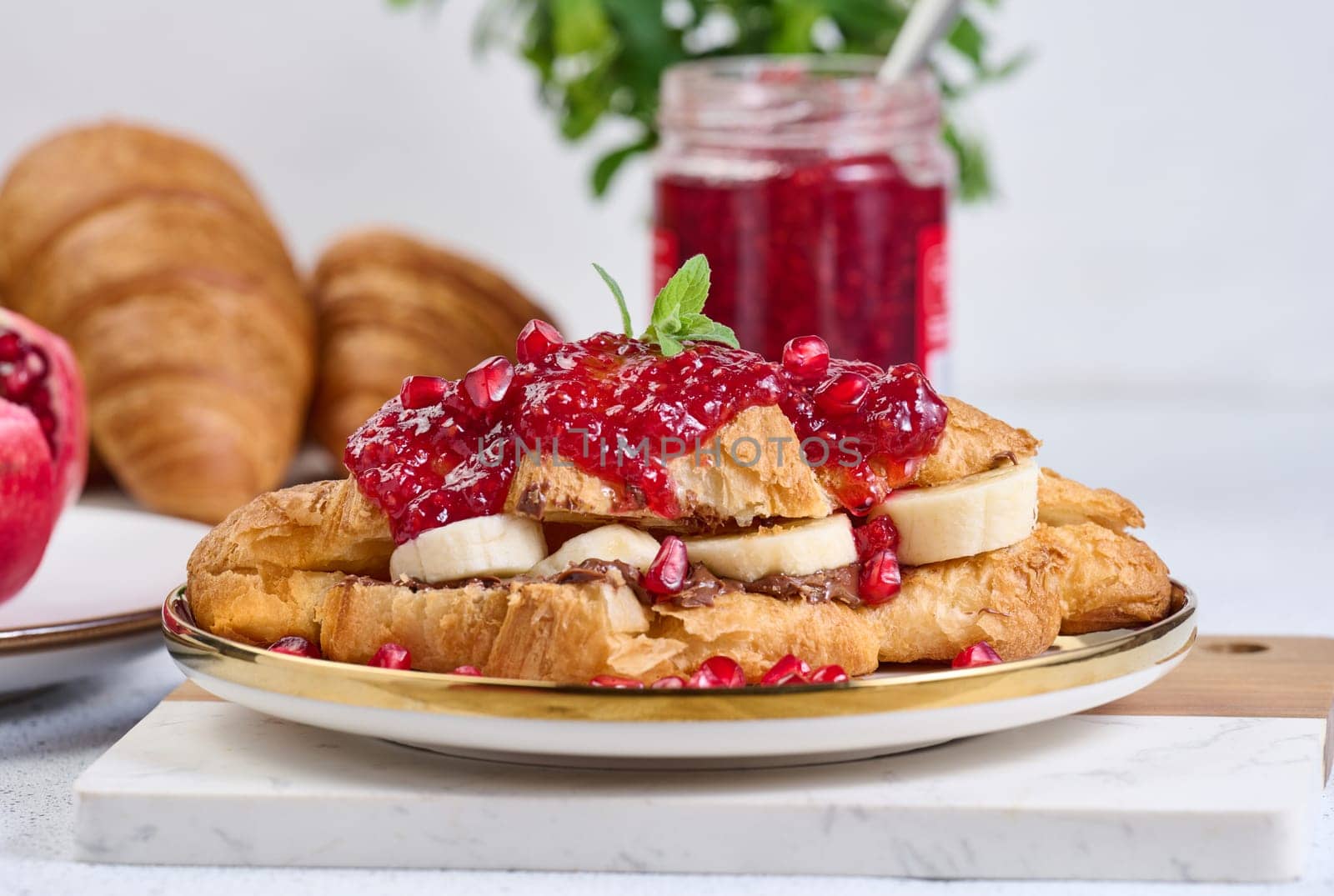 Croissant with banana pieces, chocolate paste and pomegranate seeds in a round white plate