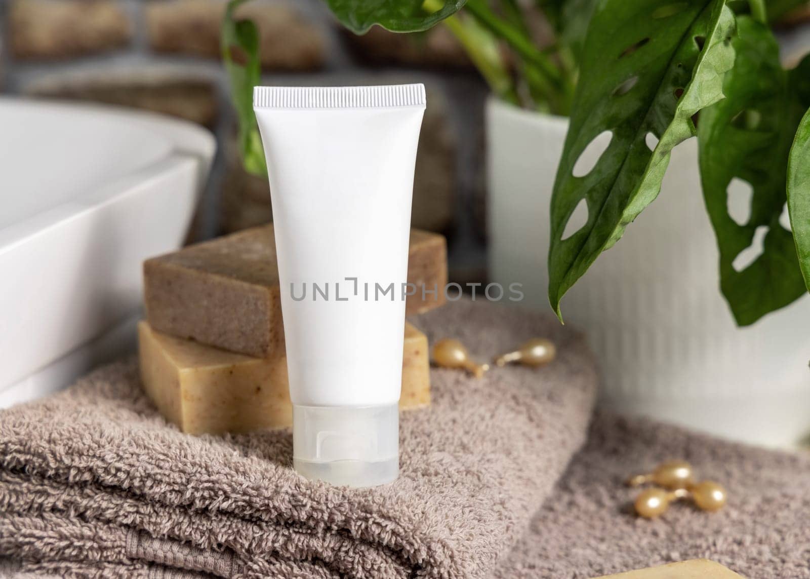 White cream squeeze tube on brown folded towels near vessel sink, soap bars and green plant in bathroom, close up, packaging mockup. Natural cosmetic product for skin care