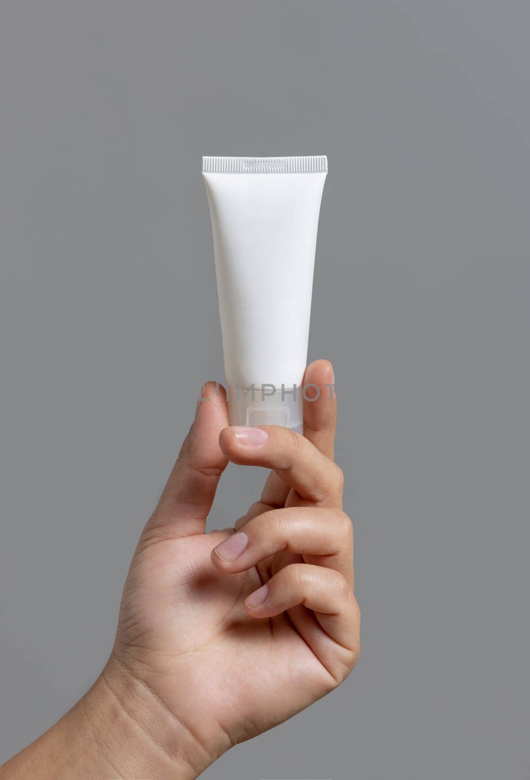 Female hand holding white squeeze cream tube against grey close up, cosmetic mockup, copy space. Skincare beauty product package, minimal composition