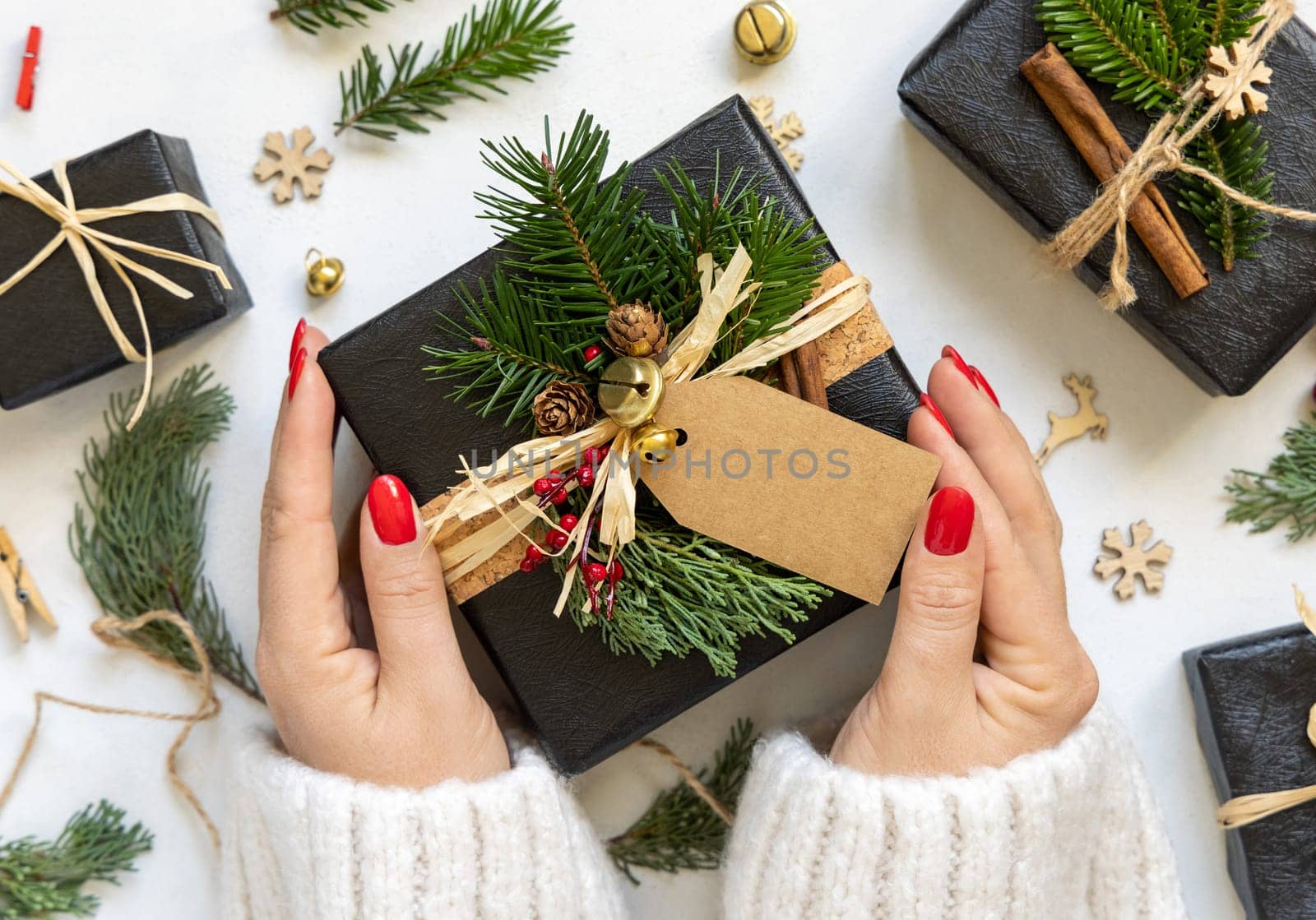 Hands with Black present decorated with fir tree branches, spices and berries, top view on white table. Winter flat lay with horizontal blank gift tag, Mockup, copy space