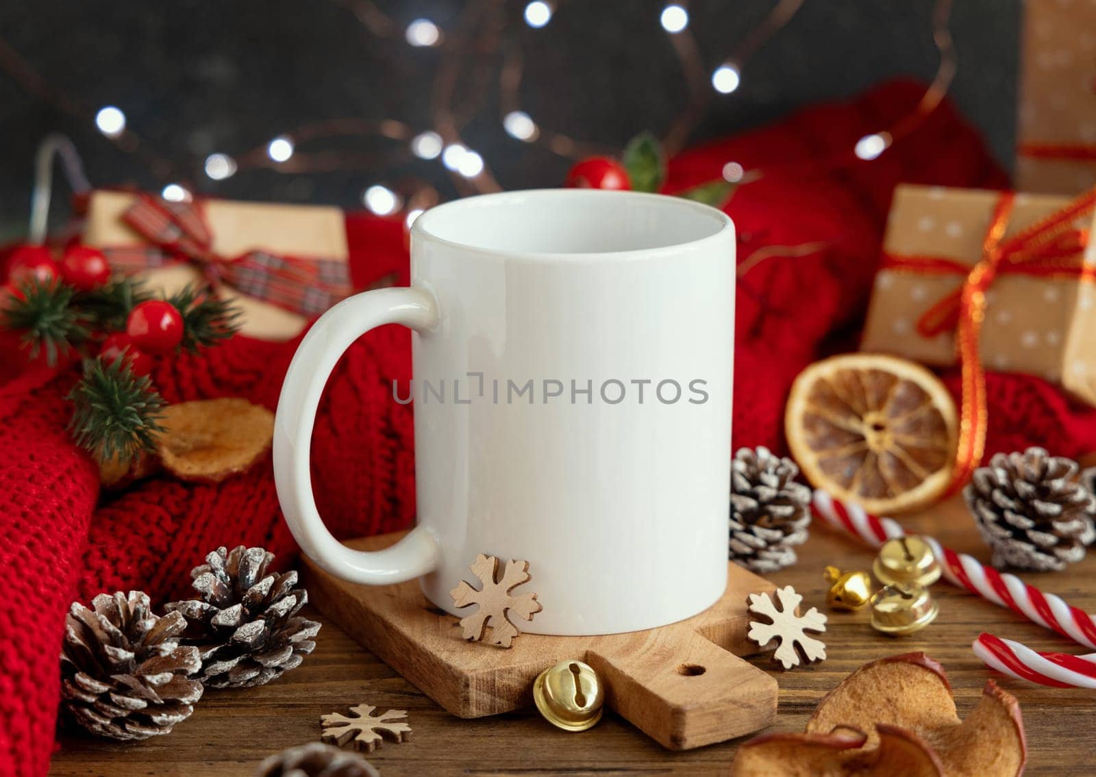 White blank coffee mug near red knitted sweater and Christmas decor against lights and wrapped presents, copy space. Warm winter family atmospheric mock up, Holiday time
