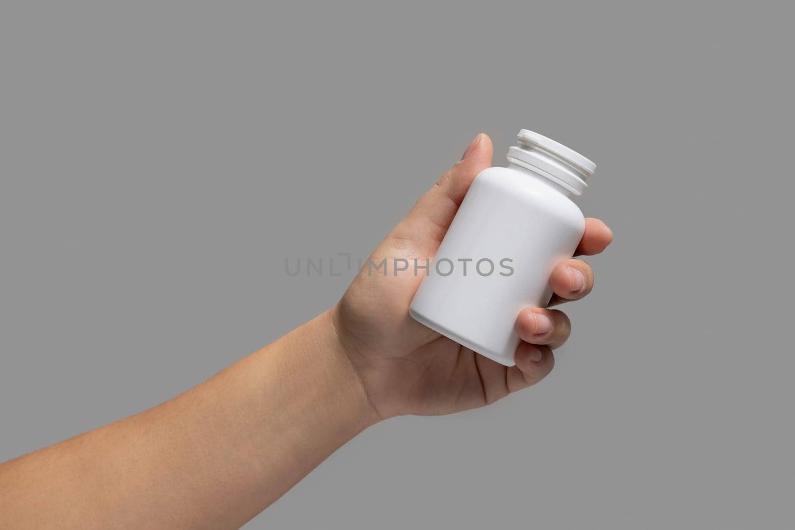 Female hand holding a white plastic pill bottle closeup against grey background. Packaging mockup for your pills or capsules herbal supplement