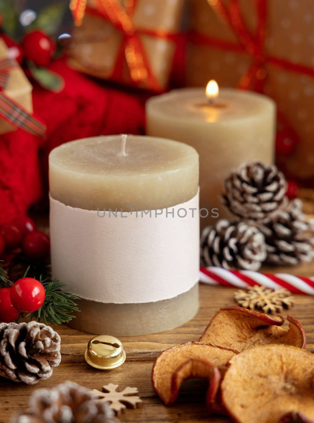 Pillar candles with blank label near red knitted sweater and Christmas decor against lights and wrapped presents, copy space. Warm winter family atmospheric mock up, Holiday time