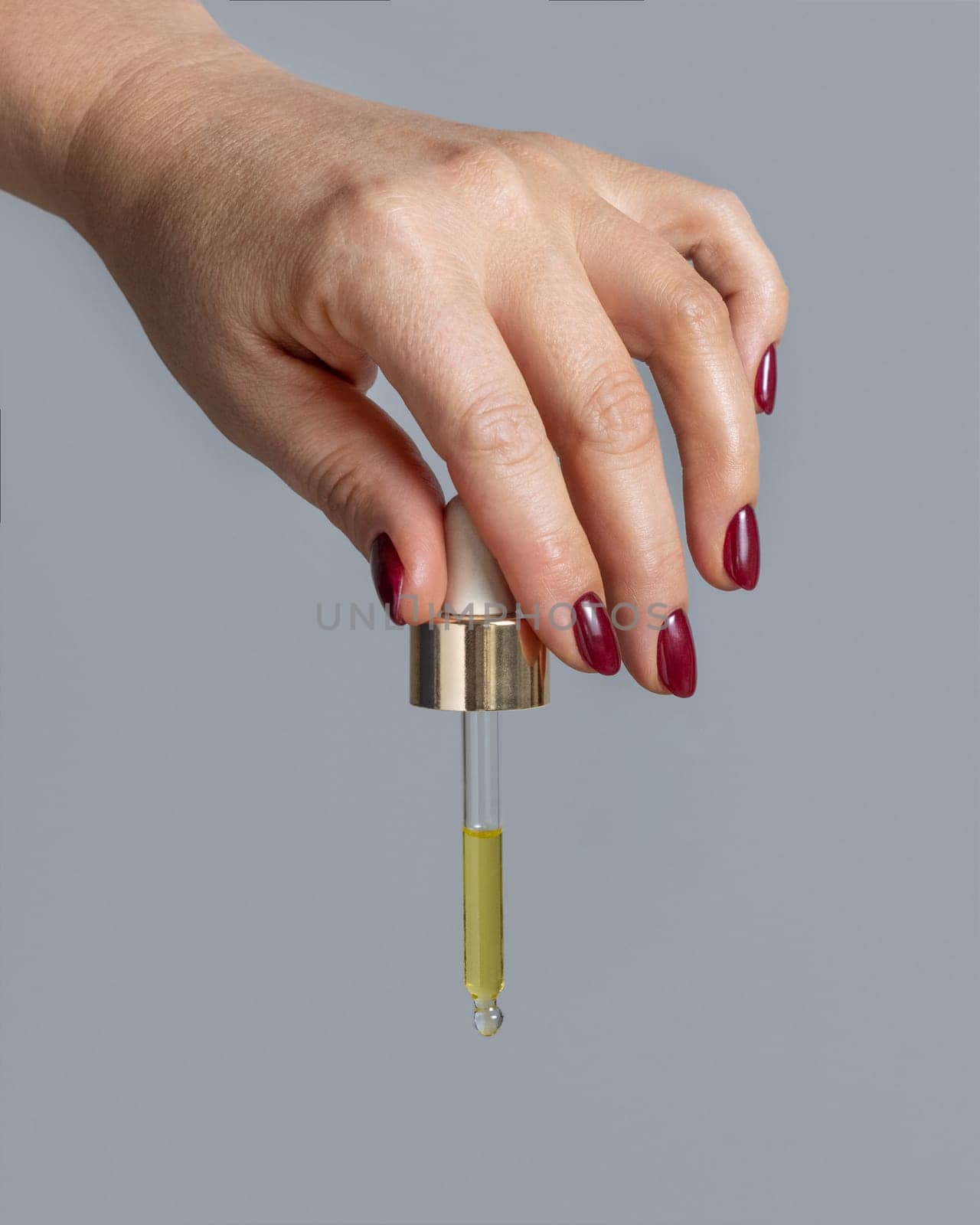 Female Hand with dark red nails holding with pipette filled with yellow liquid over grey close up. Natural skincare product, serum or essential oil. Medical research