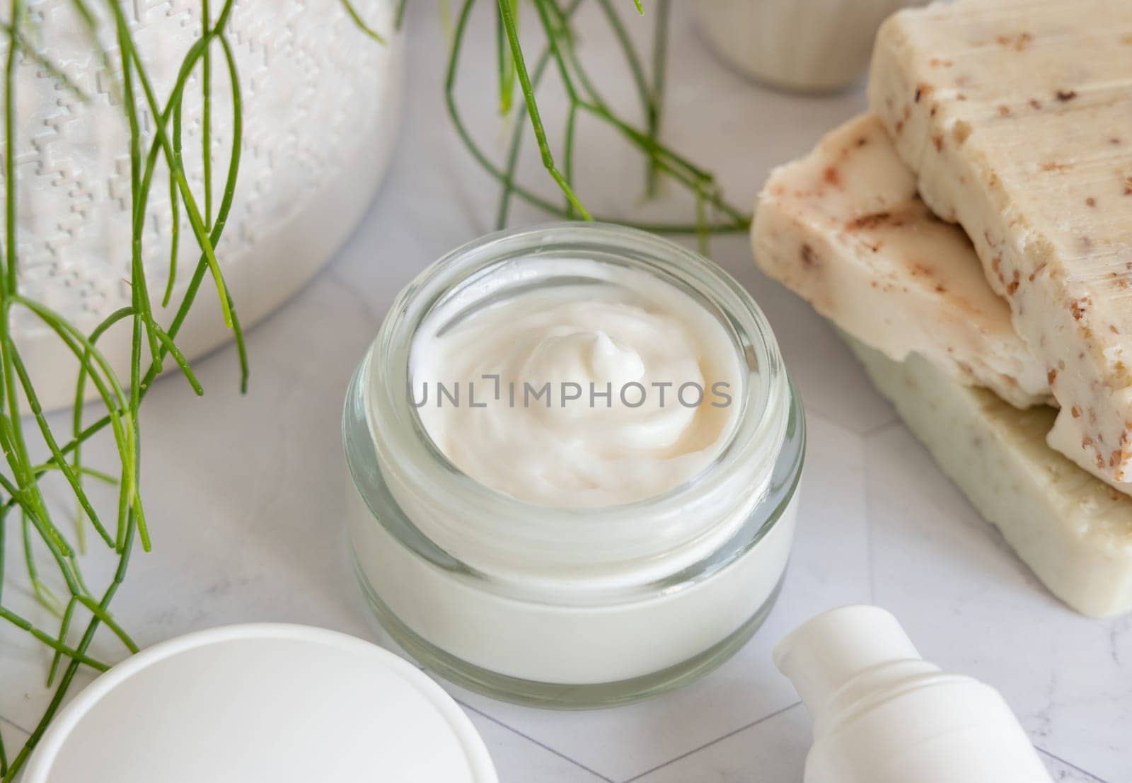 Opened cream jar with white blank lid on white marble against potted green plant and soap bars in bathroom, close up, packaging mockup. Cosmetic product for facecare, beauty routine