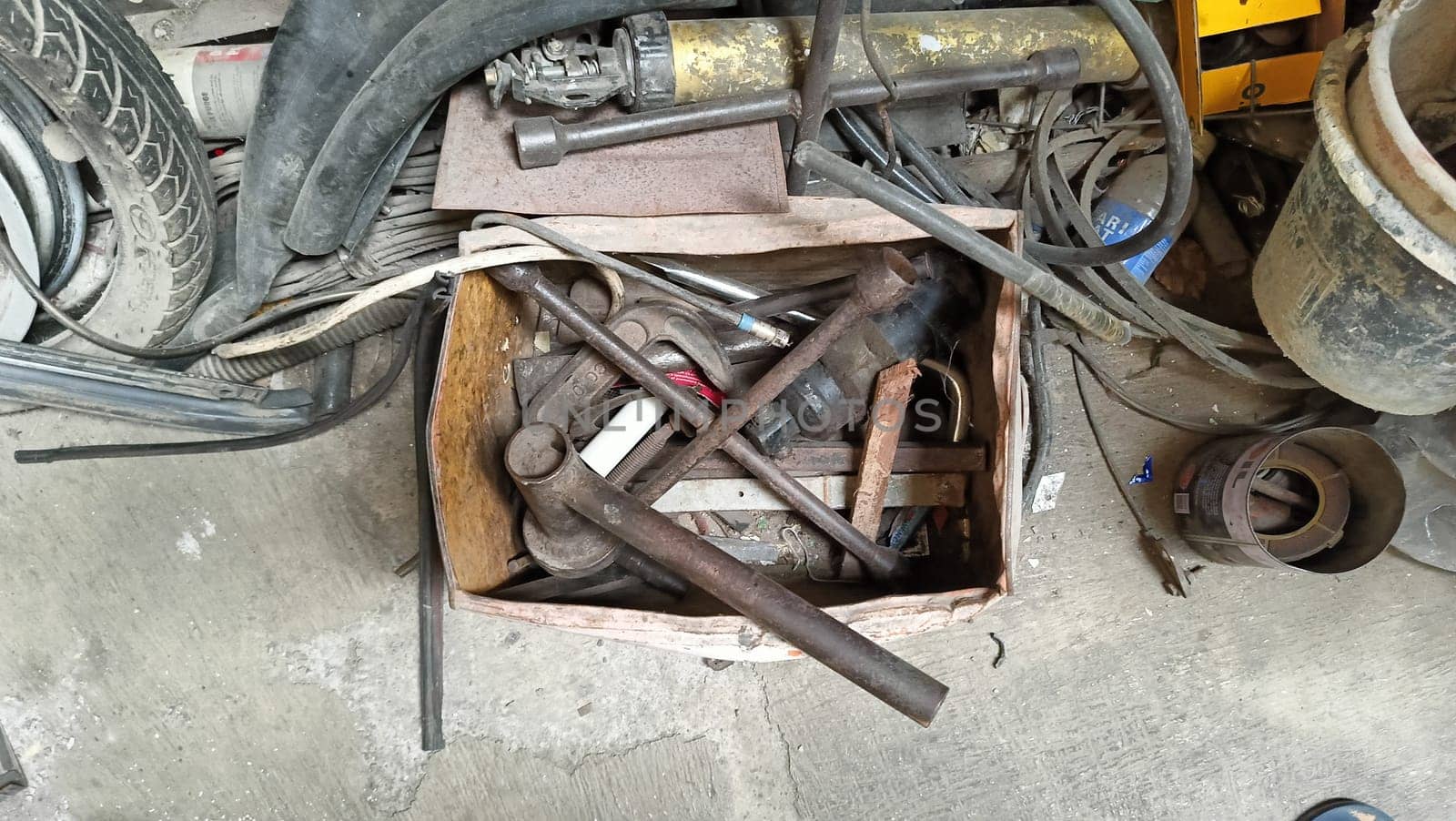 An assortment of old, rusty tools stored in a cardboard box. The scene captures the vintage, industrial feel of a workshop. A mix of wrenches, metal parts, and pipes is visible.