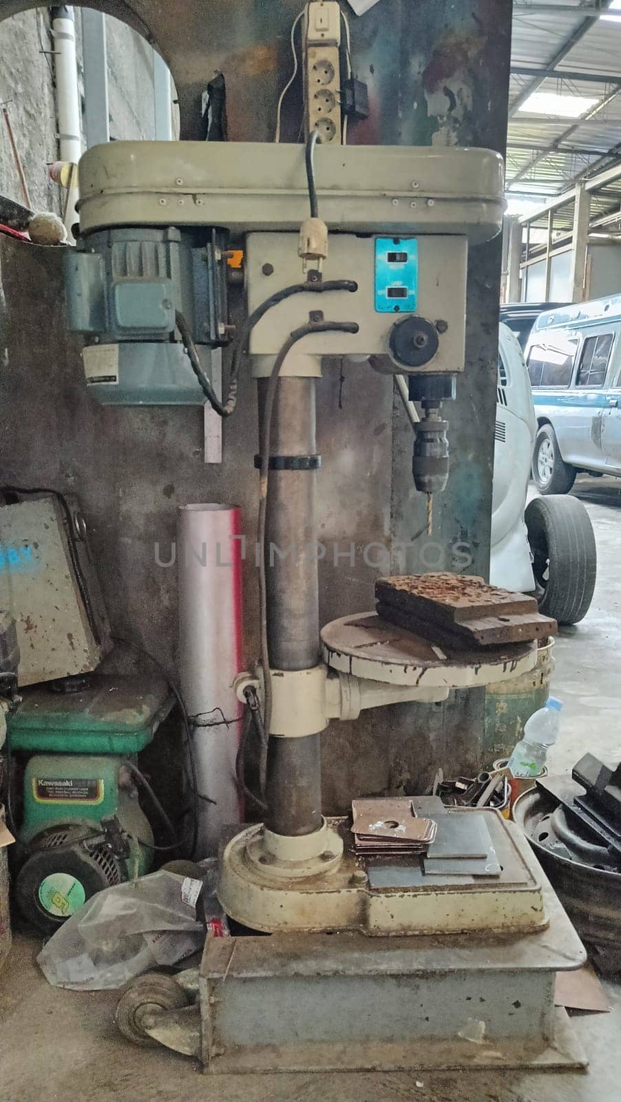 A vintage drill press stands in a cluttered workshop. The machinery, surrounded by tools and equipment, embodies industrial craftsmanship and age. This image captures the essence of a working environment.