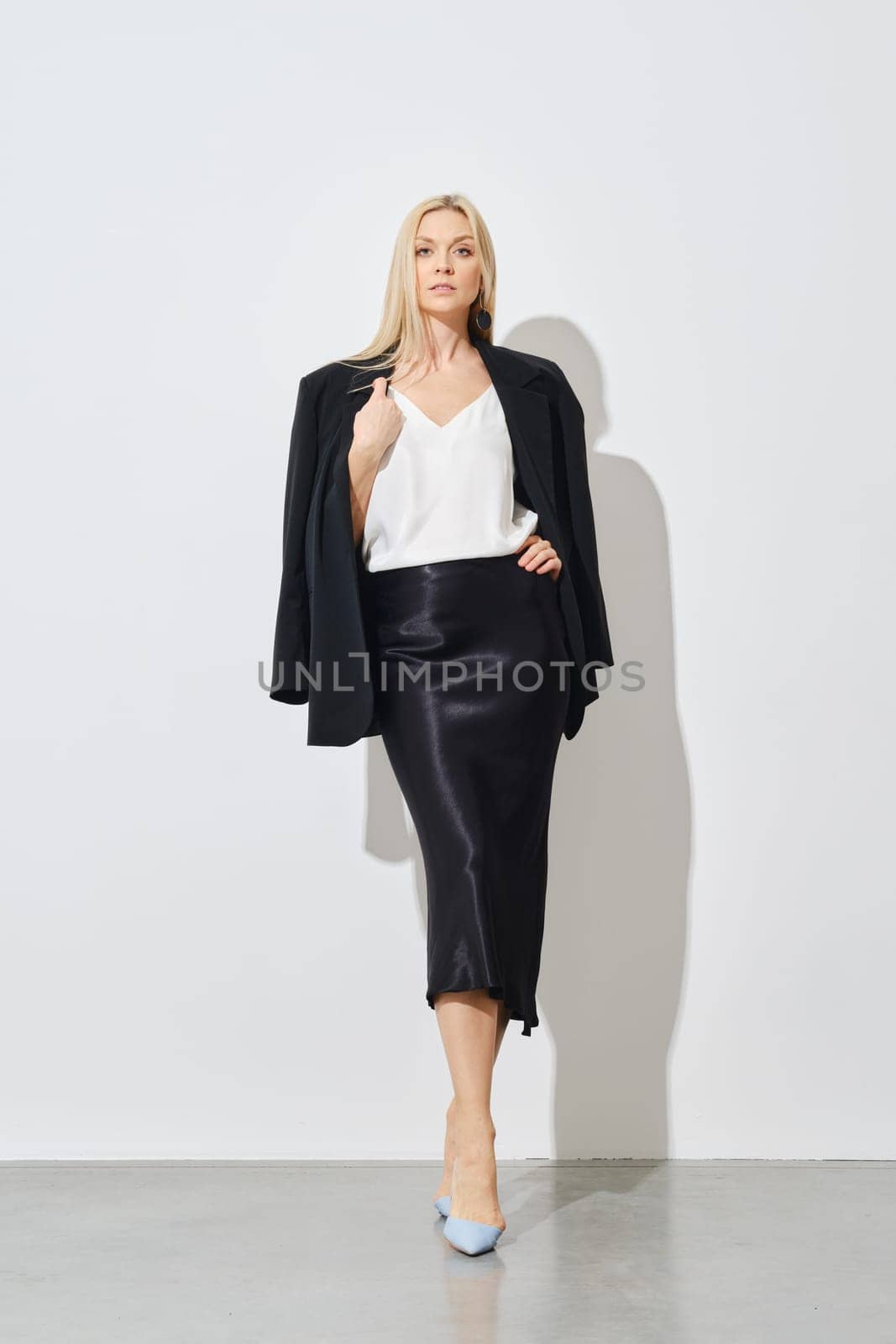 Stylish woman posing against white wall in a white blouse, a silk long skirt and a jacket draped over her shoulders