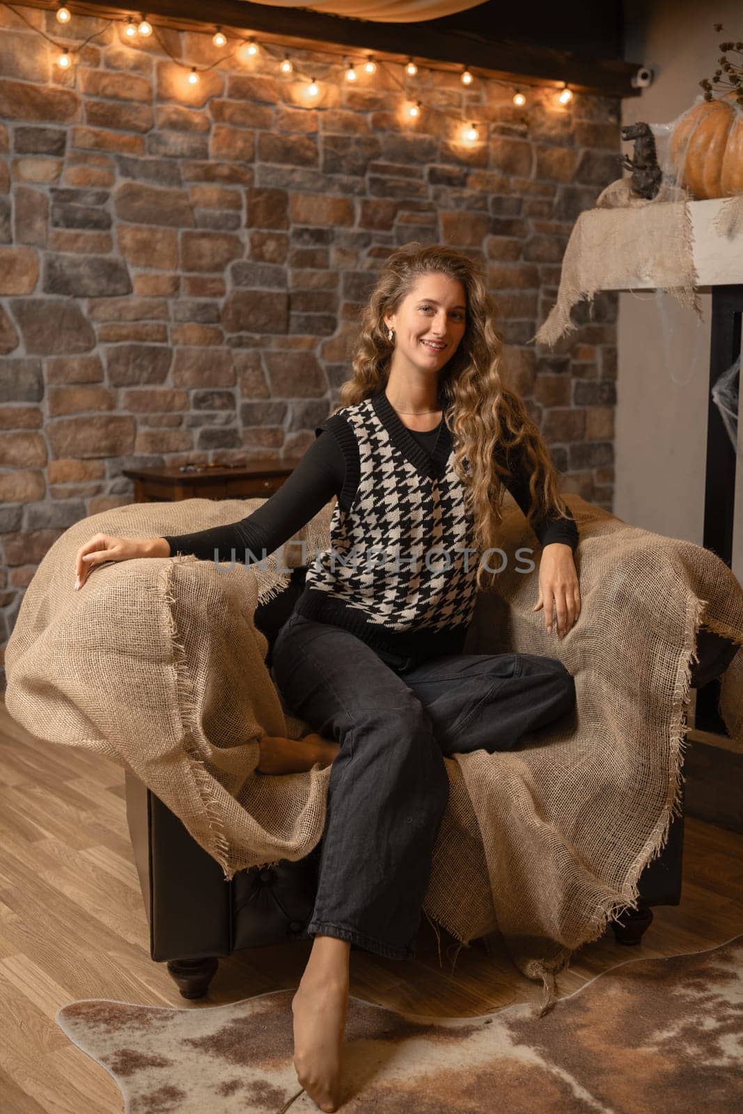 A woman sits comfortably on a couch draped with burlap in a cozy room decorated for autumn. Soft lighting casts a warm glow, creating a relaxed atmosphere perfect for unwinding.