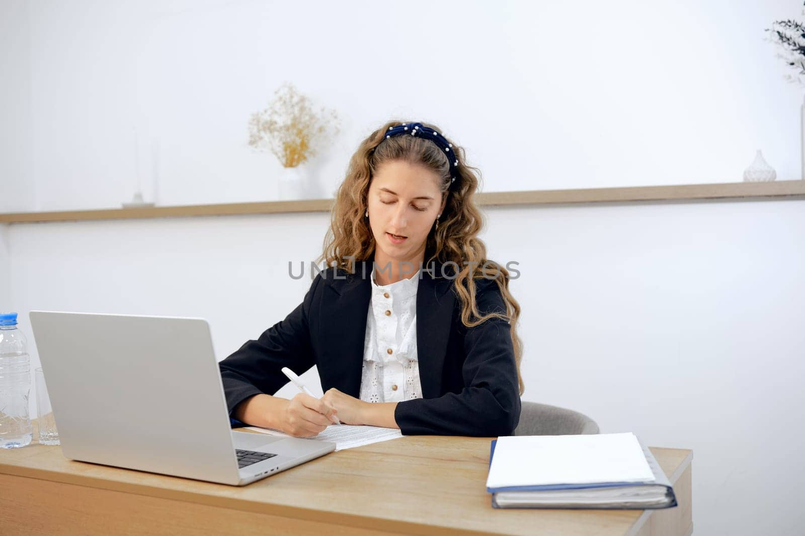A young woman sits attentively at her modern office desk, writing notes and reviewing a laptop screen. Natural light fills the space as she engages in her work with determination and concentration.
