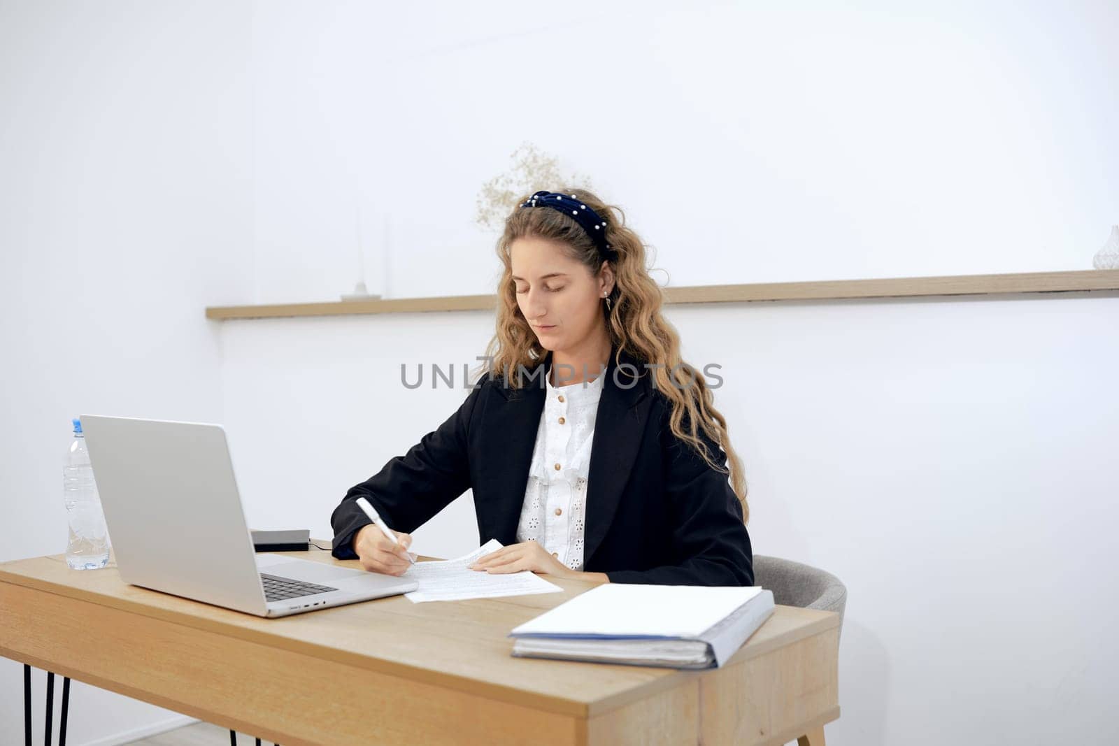 In a bright office, a young woman focuses on writing notes at her desk while a laptop is open nearby. A water bottle sits next to her as she prepares for an important task or meeting.