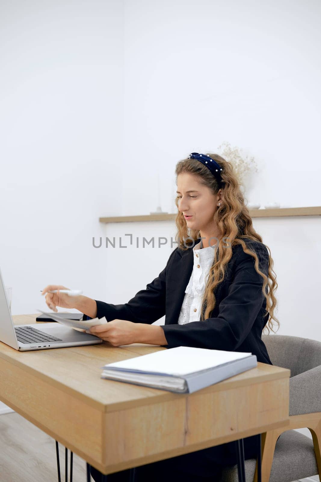 A focused young woman with curly hair sits at a minimalist desk, reviewing paperwork and typing on her laptop. The bright, neutral-colored room emphasizes her concentration on the tasks at hand.