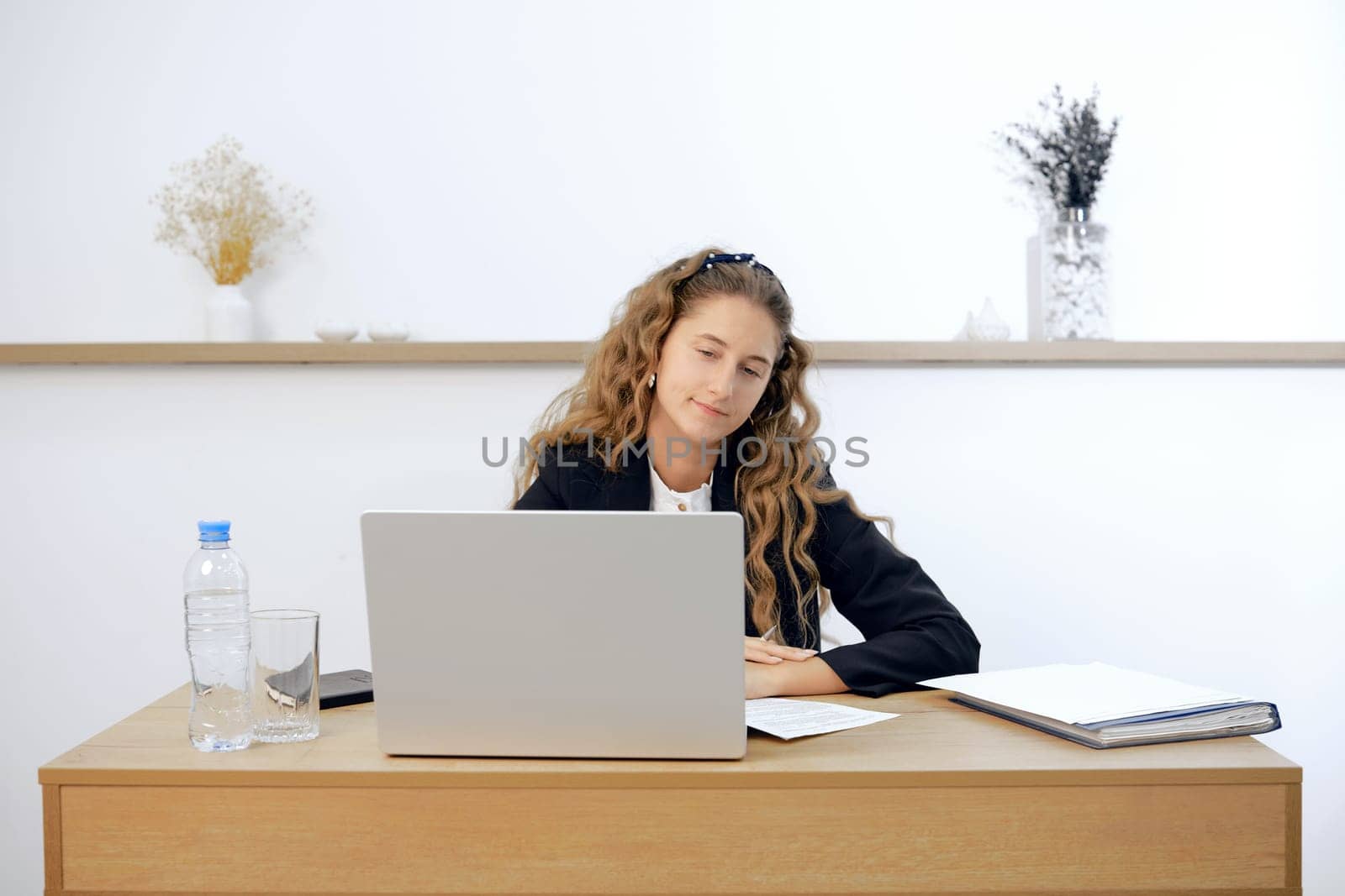 A young woman sits at a contemporary wooden desk, engaged in work on her laptop. Papers and a water bottle are placed nearby, creating a tidy workspace in a bright, minimalist office environment.