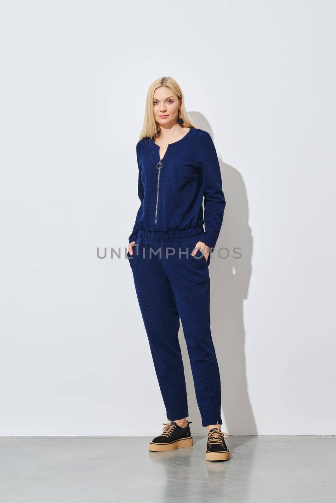 Blonde woman posing in knitted blue pantsuit against white wall