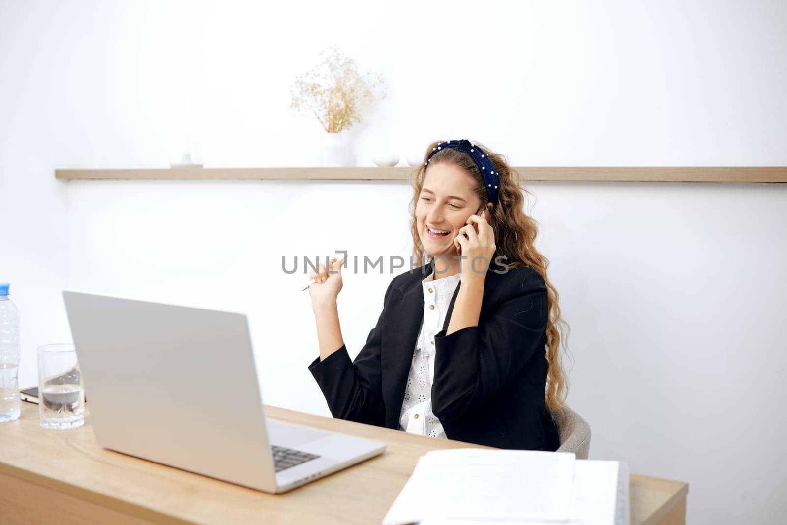 A young woman with long, curly hair smiles during a phone call and takes notes at her tidy desk. The bright office features light wood accents and minimal decor, creating an inviting workspace.