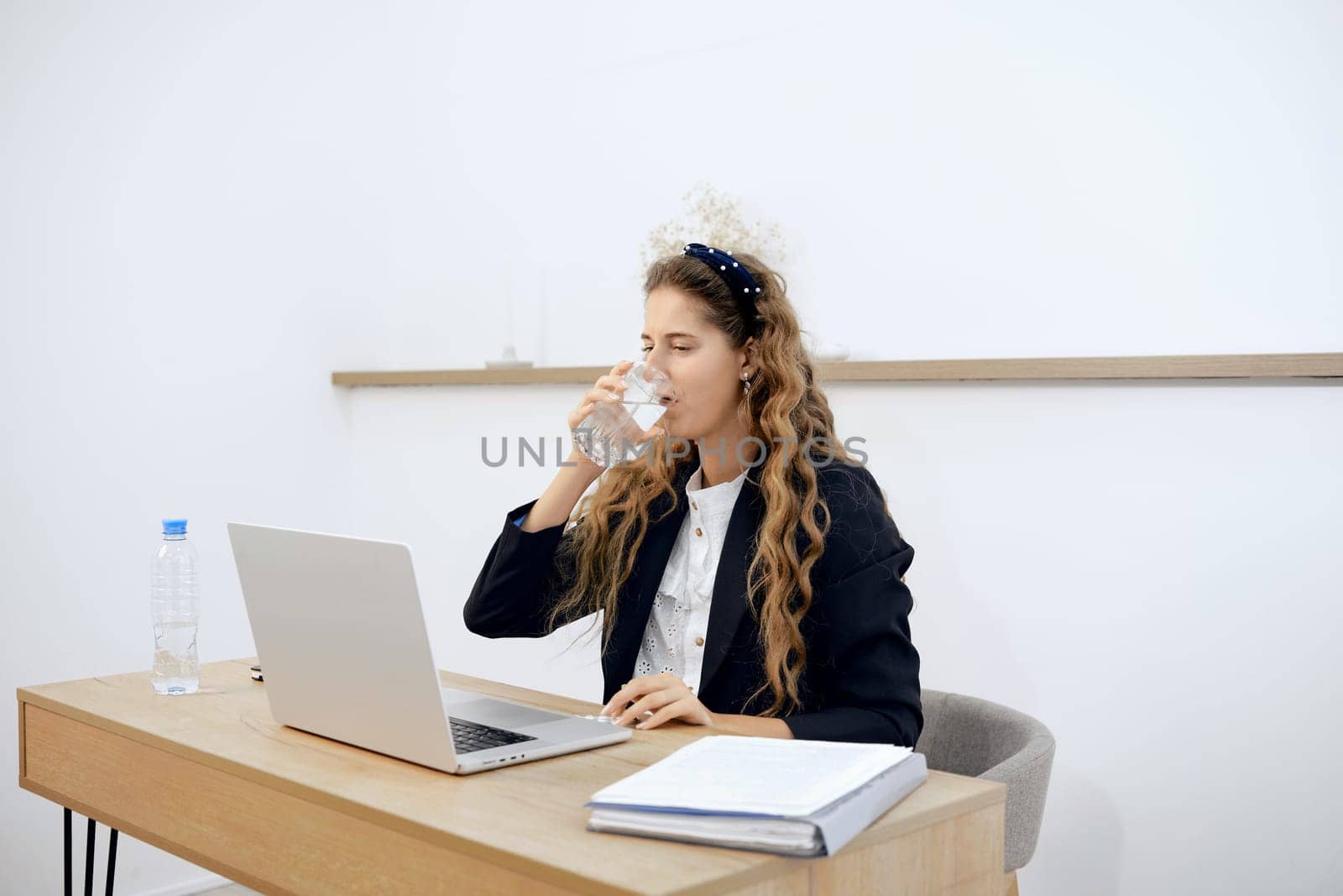 A professional woman with curly hair sits at a desk, sipping water from a glass while focused on her laptop.
