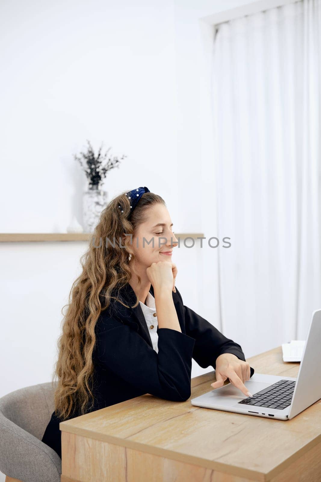 A woman with wavy hair sits at a wooden desk, focused on her laptop. She wears a black blazer and appears engaged in her work. The room is well-lit with white curtains and simple decor.