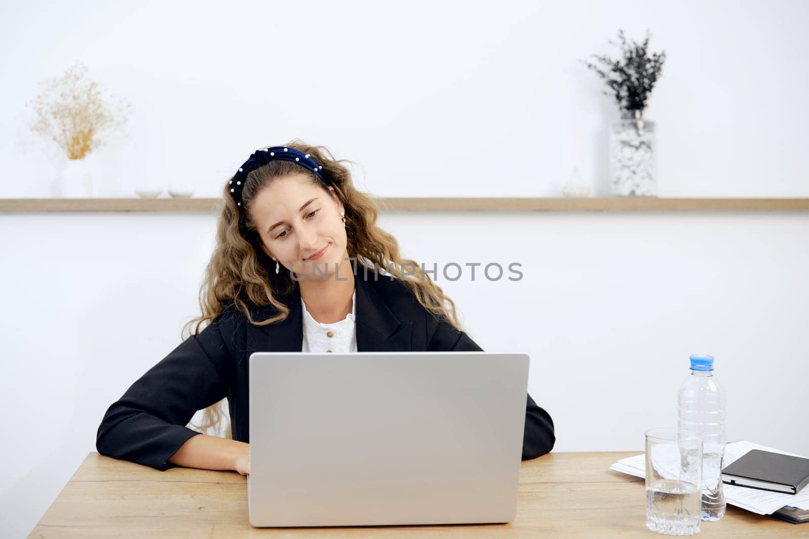 A focused woman with curly hair sits at a desk, engaged with her laptop. The workspace is minimal, with a bottle of water and notebook nearby, creating a productive and calm atmosphere.